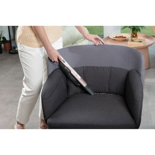A person is using a BLACK+DECKER handheld vacuum cleaner to clean crumbs from a dark-colored armchair in a modern living room.