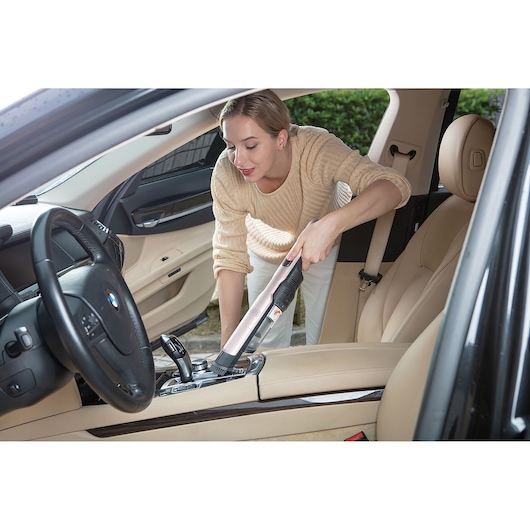 A person is using a BLACK+DECKER handheld vacuum cleaner to clean the interior of a beige-colored car, focusing on the area near the center console.