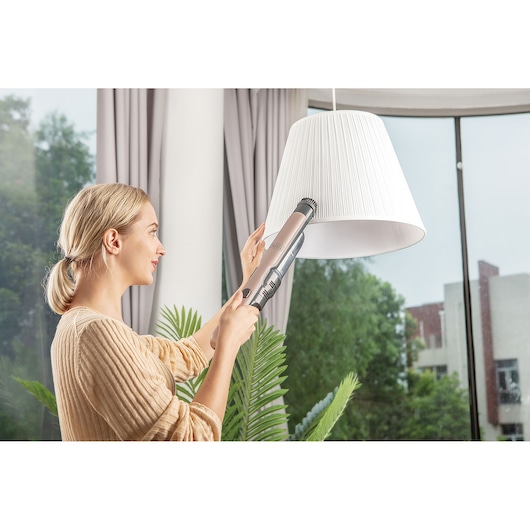 A person using a BLACK+DECKER handheld vacuum cleaner to clean a white lampshade in a bright room with large windows and plants. The person's face is blurred for privacy.