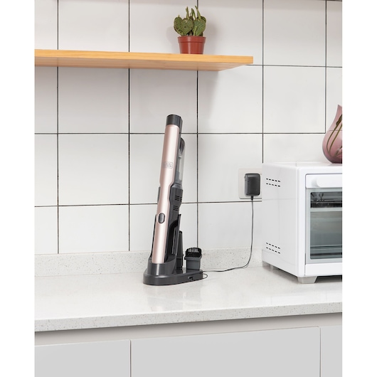 A BLACK+DECKER handheld vacuum cleaner in a charging dock on a kitchen countertop. The device is plugged into a wall socket, and next to it is a white appliance, likely a microwave oven. There are tiled walls, a wooden shelf with a small potted cactus, and some pink decor visible.