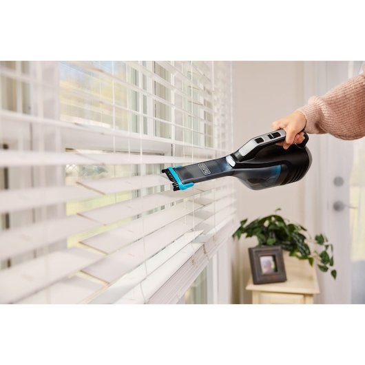 BLACKDECKER DVD320B handheld vacuum cleaning window blinds with brush attachment indoors.