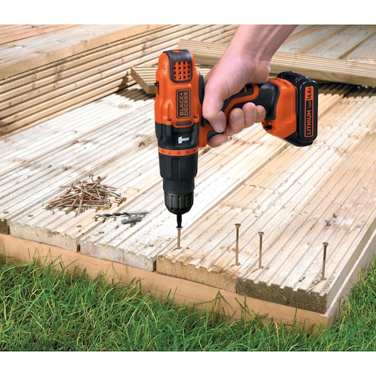 A person using a BLACK+DECKER cordless drill to drive a screw into a wooden deck. The drill is orange and black and marked with 'Lithium 14.4V'. Several screws and drill bits are visible on the wooden surface.