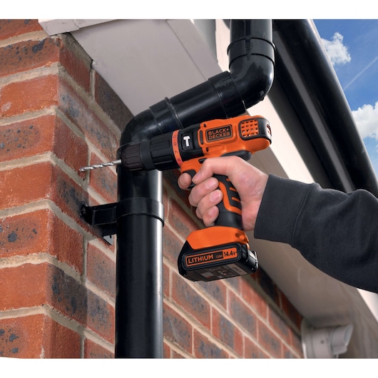 A person is using a BLACK+DECKER 14.4V lithium cordless drill to drill into a brick wall near a black gutter pipe.