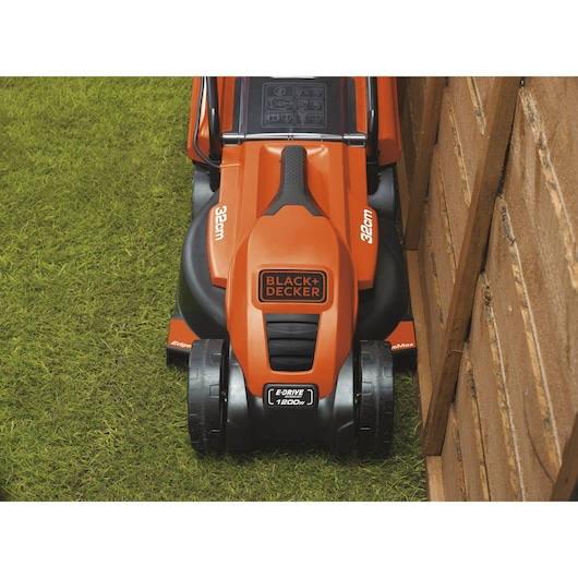Photo of BLACK+DECKER EMAX32S orange electric lawn mower cutting grass close to a wooden fence, top view.
