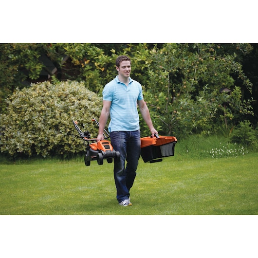 Person carrying BLACKDECKER EMAX32S orange lawn mower and grass box on grass in garden.