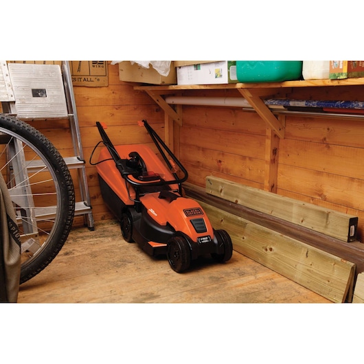 BLACK+DECKER EMAX32S orange lawn mower in a wooden shed, shown from the side among tools and wood planks.