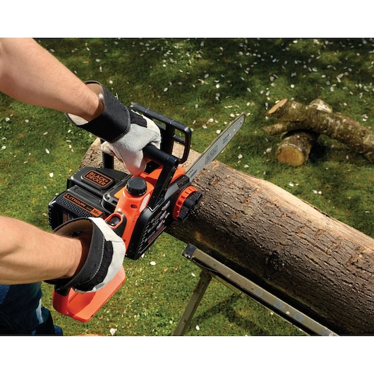 A person wearing gloves is using a BLACK+DECKER cordless chainsaw to cut a log placed on a metal support in a grassy outdoor setting.