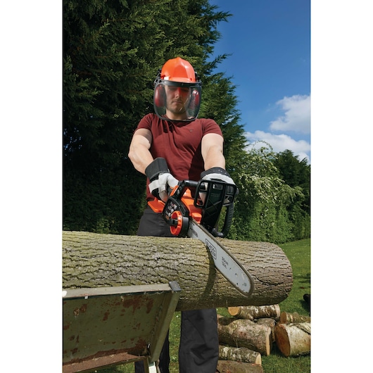 A person wearing protective gear uses a BLACK+DECKER GKC3630L20 cordless chainsaw to cut a large log outdoors. The scene shows safety equipment including gloves, helmet, face shield, and earmuffs, with logs and greenery in the background.