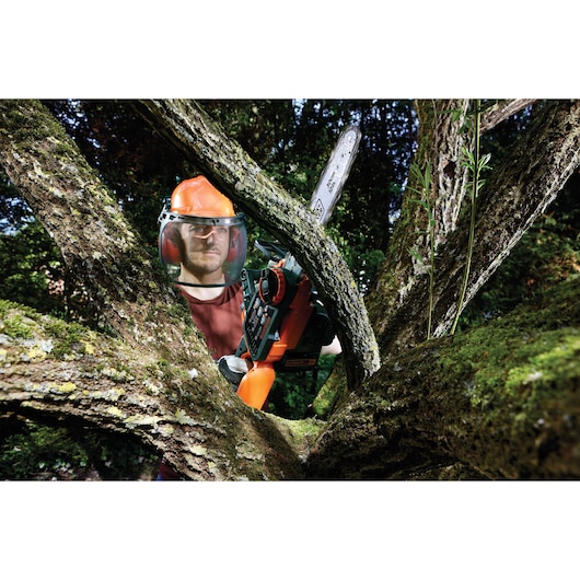 Person wearing protective gear using a BLACK+DECKER chainsaw to cut tree branches outdoors. The chainsaw model is GKC3630L20.