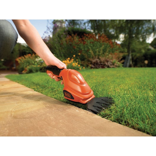 A person is using a BLACK+DECKER cordless grass shear to trim the edge of a lawn next to a pavement, with a garden and flowers visible in the background.