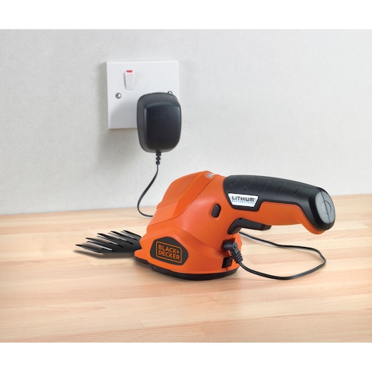 A BLACK+DECKER lithium battery-powered handheld garden shear is being charged with a power adapter plugged into a wall socket. The device is placed on a light wooden surface.