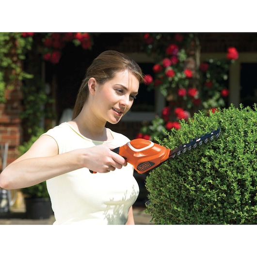 A person is using a BLACK+DECKER GSL700 cordless shrub shear to trim a bush in a garden. The tool is orange and black, and the setting includes flowers and greenery.