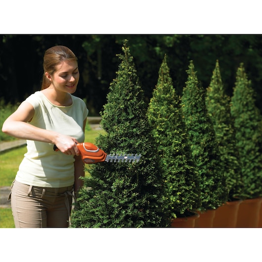 A person with a blurred face is using a BLACK+DECKER GSL700 cordless hedge trimmer to trim a bush shaped like a cone. Several similar potted bushes are lined up in the background.