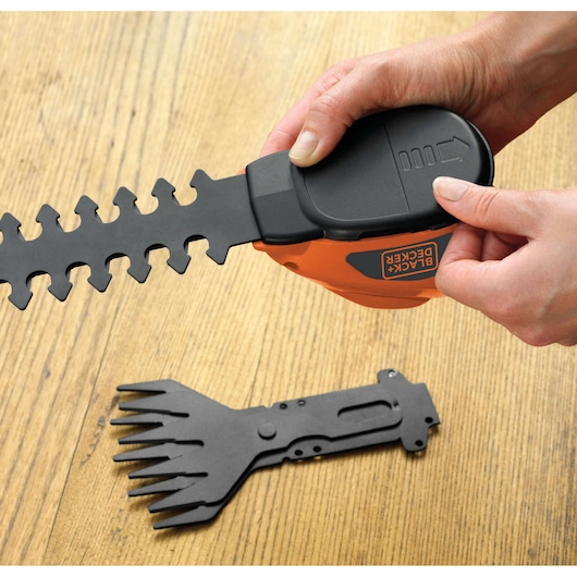 A close-up view of hands attaching a blade to a BLACK+DECKER GSL700 garden tool. Another interchangeable blade is placed on a wooden surface.