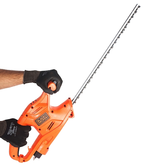 A person wearing black gloves is holding an orange BLACK+DECKER electric hedge trimmer with a long blade against a white background.