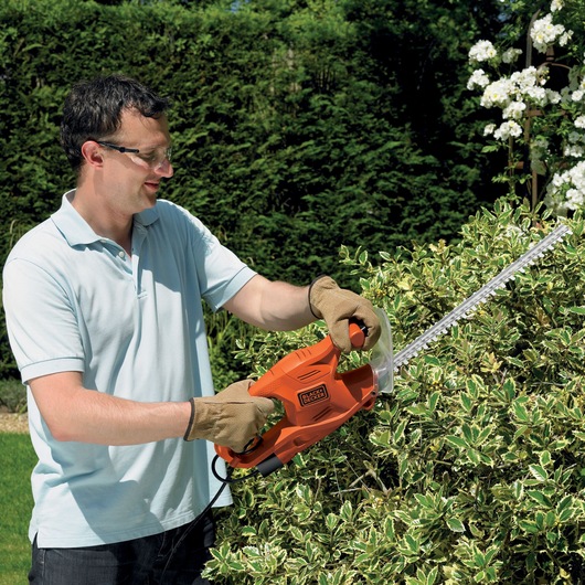 A person wearing gloves is using an orange BLACK+DECKER electric hedge trimmer to cut bushes in a garden.