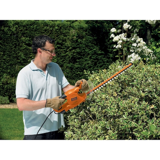 A person using a BLACK+DECKER GT4550 electric hedge trimmer to trim bushes in a garden. The person is wearing gloves and a light blue polo shirt, with their face blurred for privacy.