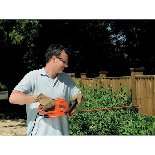 Person trimming hedge with BLACK+DECKER GT5055 orange electric hedge trimmer outdoors near wooden fence.