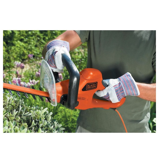 BLACK+DECKER GT6030 hedge trimmer in use, person wearing gloves trimming green bushes outdoors.