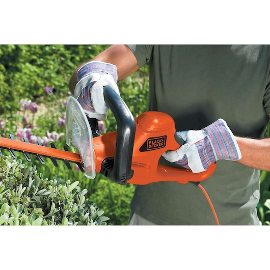 A person wearing protective gloves is using an orange BLACK+DECKER electric hedge trimmer to trim bushes in a garden.