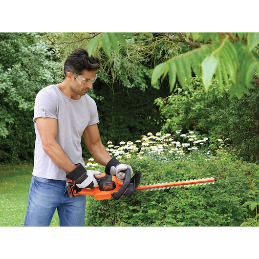 Photo of person using BLACK+DECKER GTC18452PC hedge trimmer outdoors, trimming green bushes in a garden.