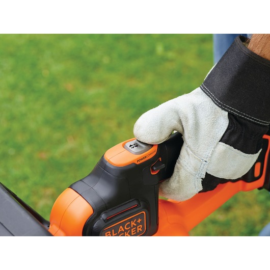 A gloved hand operating a BLACK+DECKER cordless hedge trimmer outdoors on a grassy lawn. The person is pressing a button labeled 'PowerCommand' on the tool.