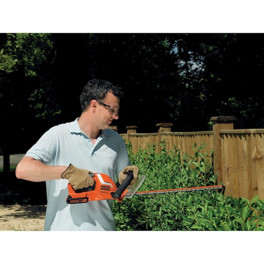 A person wearing gloves uses a BLACK+DECKER hedge trimmer (model GTC1850L20) to trim bushes near a wooden fence in a garden.