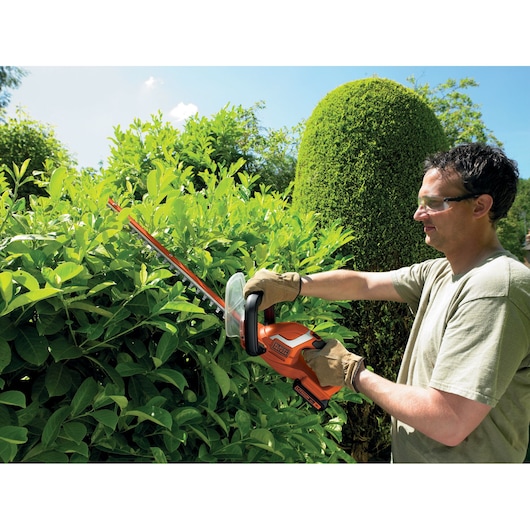 A person wearing gloves is trimming hedge bushes with an orange Black+Decker cordless hedge trimmer outdoors. The person's face is blurred for privacy.
