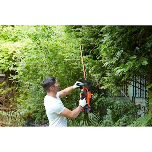 A person wearing gloves uses a BLACK+DECKER cordless hedge trimmer to trim green hedges in a garden.