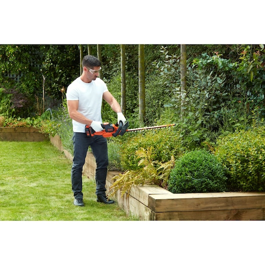 A person wearing gloves is using a BLACK+DECKER cordless hedge trimmer to cut shrubs in a well-maintained garden with raised wooden beds.
