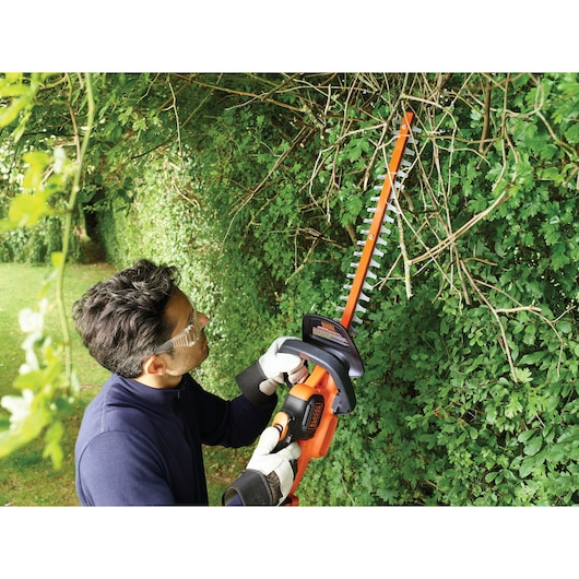 A person wearing gloves uses a BLACK+DECKER cordless hedge trimmer (model GTC36552PC) to trim thick branches and hedges in a garden. The person's face is blurred for privacy.