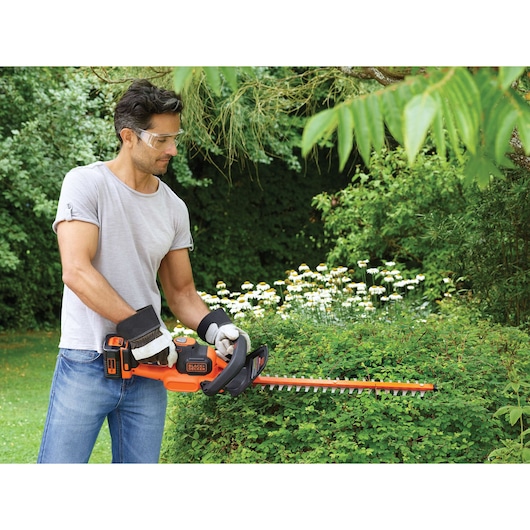 A person using a BLACK+DECKER cordless hedge trimmer to trim a bush in a garden, wearing gloves and a grey t-shirt, surrounded by green foliage and flowers.