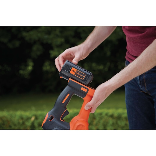 A person inserts a BLACK+DECKER 36V lithium battery into an orange and black cordless tool, likely a hedge trimmer, in an outdoor garden setting.