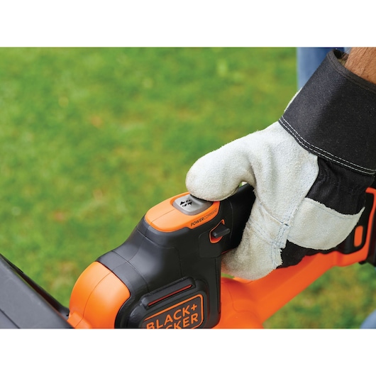 Close-up of a gloved hand operating the POWERCOMMAND button on a BLACK+DECKER garden tool with orange and black casing, on a grassy outdoor background.