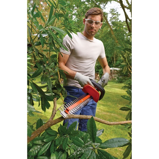 Person using BLACKDECKER GTC5455PC hedge trimmer to cut a tree branch outdoors, wearing gloves in a garden.