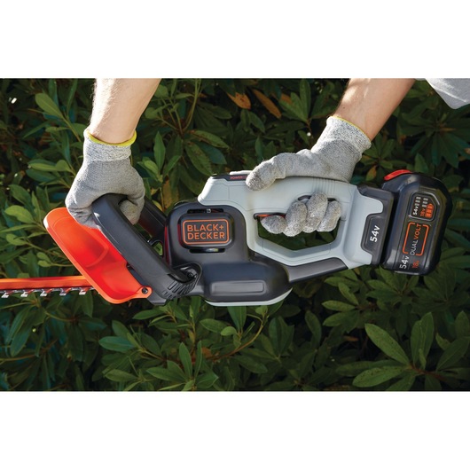 Photo of BLACK+DECKER GTC5455PC hedge trimmer with 54V battery held by gloved hands in front of green bushes.
