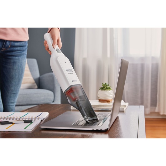 BLACK+DECKER HLVC315B11 handheld vacuum being used to clean a laptop keyboard on a desk in a home setting.