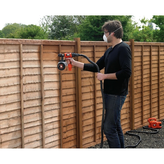 BLACKDECKER HVLP400 paint sprayer in use on wooden fence outdoors, connected by hose.
