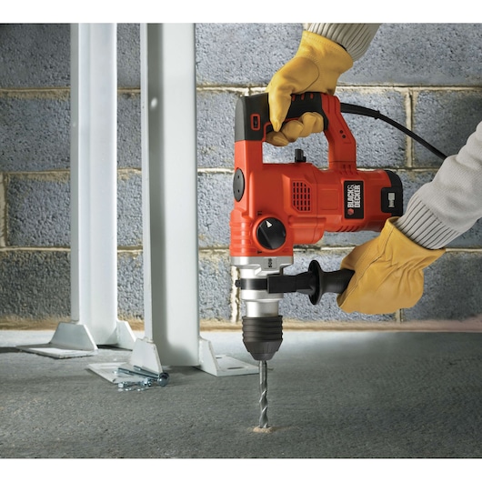 A person wearing yellow gloves is using a BLACK+DECKER KD1250K hammer drill to drill into a concrete floor near a metal structure and brick wall.