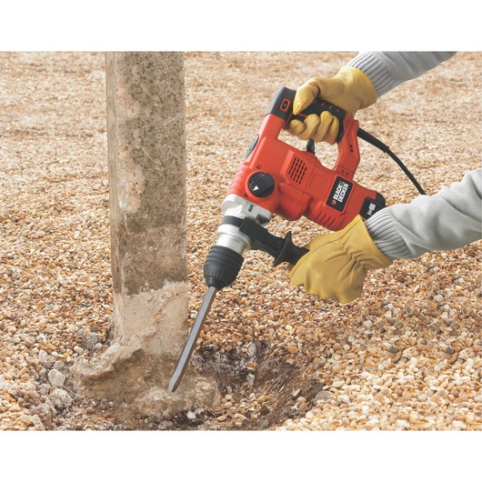 A person wearing yellow work gloves uses a BLACK+DECKER KD1250K rotary hammer drill to break concrete at the base of a post on a gravel surface.