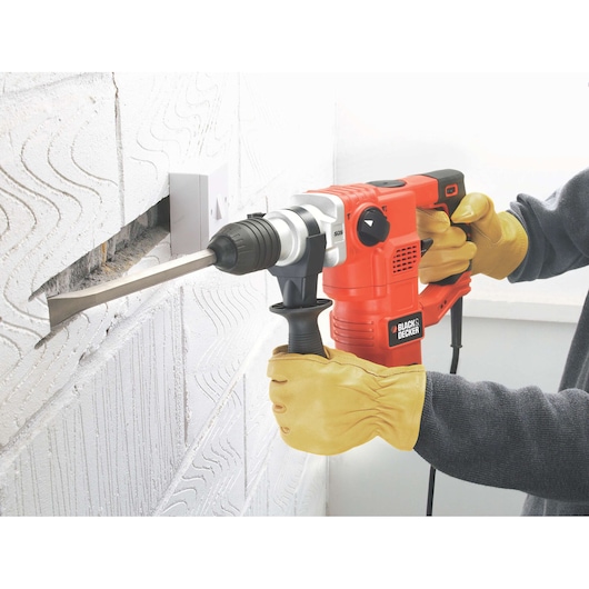 A person wearing yellow gloves uses a BLACK+DECKER KD1250K rotary hammer drill to chisel a groove into a white concrete wall.