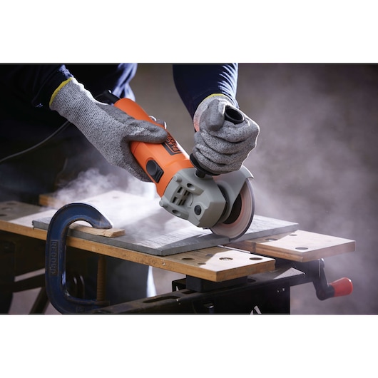 BLACKDECKER KG115 angle grinder in use cutting tile with gloves, workbench and clamp visible.