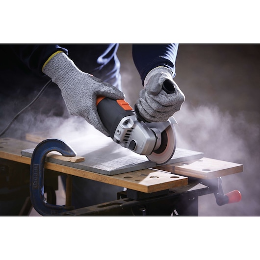 A close-up of a person wearing protective gloves using a BLACK+DECKER angle grinder to cut a piece of material fixed on a workbench, with dust visible in the air.