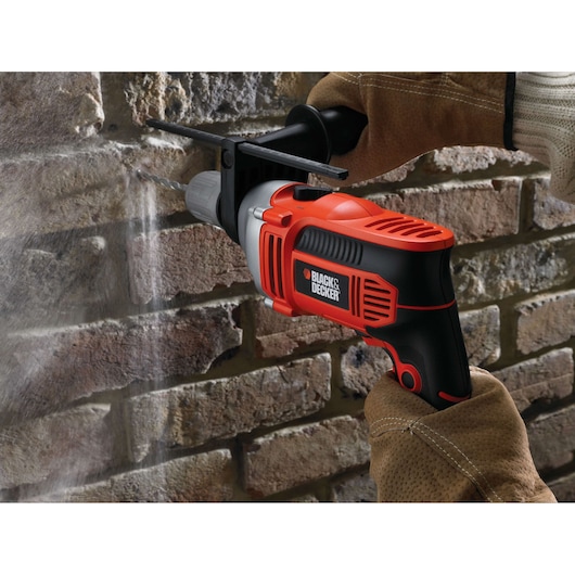 A person wearing gloves uses a BLACK+DECKER electric drill to drill into a brick wall.
