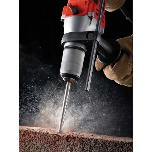 Close-up image of a power drill with a side handle being used to drill into a brick or masonry surface, creating dust and debris. The drill is held by a gloved hand.