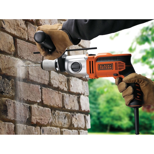 A close-up of a person wearing gloves and using a BLACK+DECKER electric drill to drill into a brick wall outdoors. Dust is visible coming from the drilled hole.