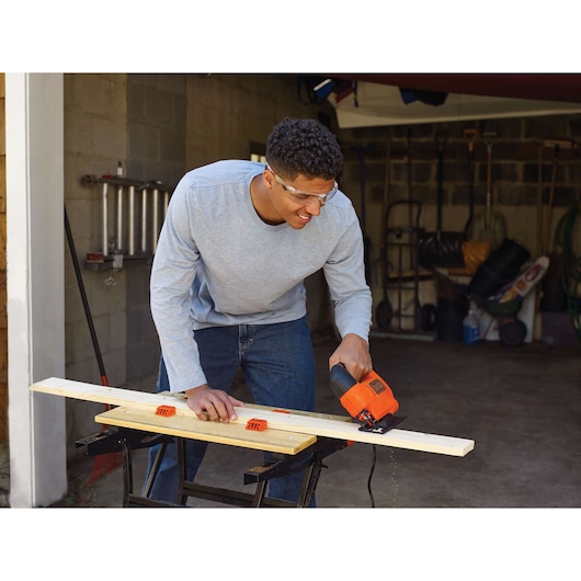 A person using a BLACK+DECKER KS701E jigsaw to cut a piece of wood, with the wood secured on a workbench in a garage or workshop setting.