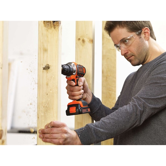 A person using a BLACK+DECKER cordless drill to bore a hole in a wooden beam. The person's face is blurred for privacy.