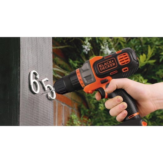 A person uses a BLACK+DECKER cordless drill to attach metal house numbers to a wooden surface outdoors. The drill is orange and black, and the environment features greenery and a brick wall in the background.