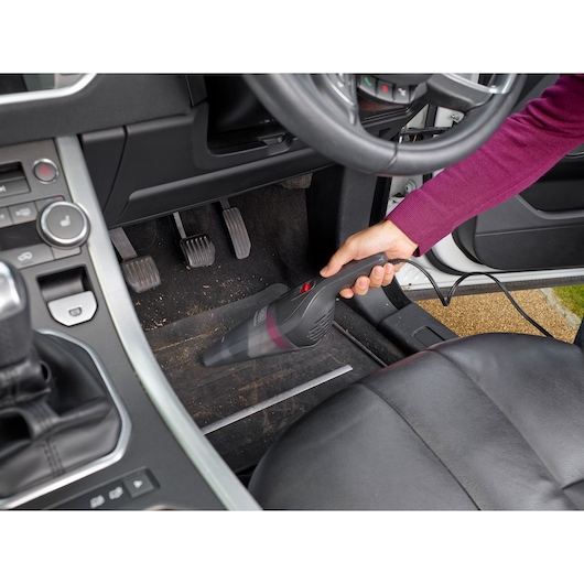 A person using a BLACK+DECKER handheld vacuum cleaner to clean the floor of a car, focusing on the area near the pedals.